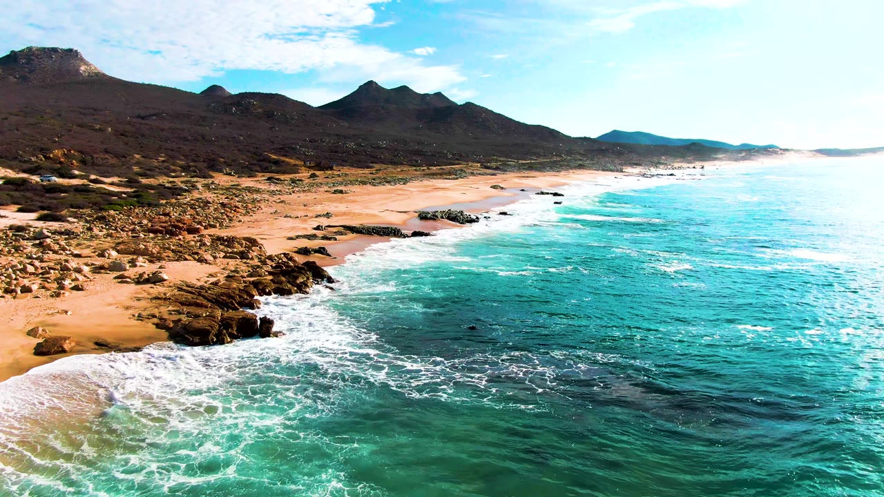 Aerial view of turquoise surf meeting a rocky sandy coast with mountains behind.