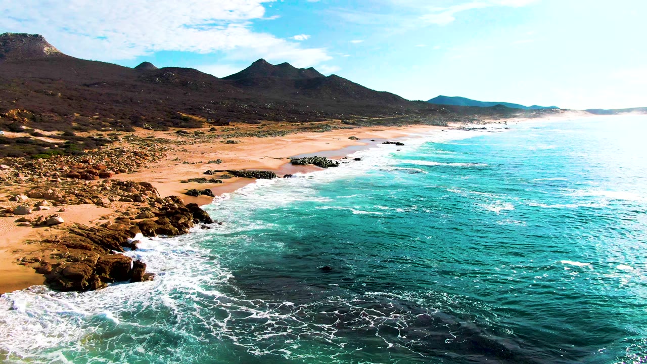 Punta Buzzards coastline with surf spreading farther across the near rocks, tan beach running diagonally, mountain ridges inland, and no visible people.
