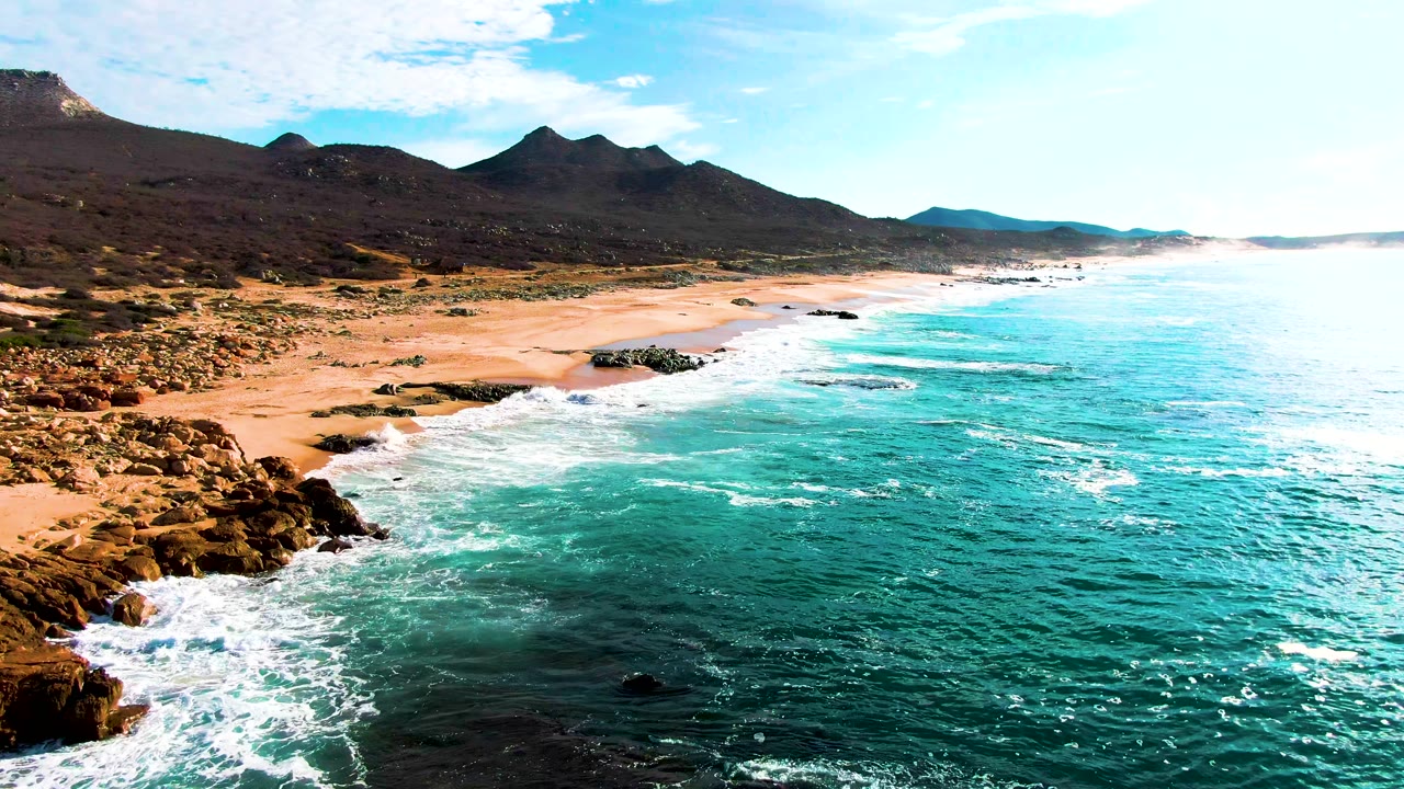 Aerial view over Punta Buzzards with the rocky shoreline shifted left in frame, bright surf bands over turquoise water, broad sandy beach, mountains, and no visible people.