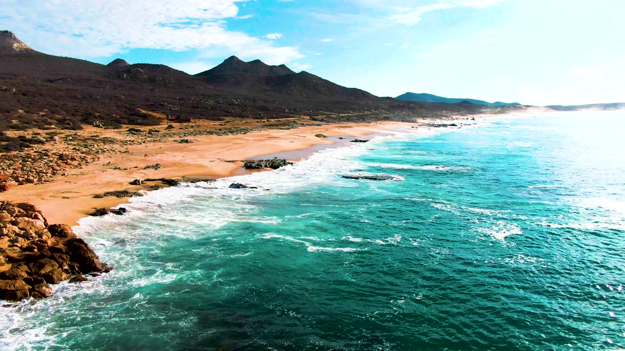 Punta Buzzards aerial frame with foreground rocks at lower left, foamy water crossing the shoreline, open turquoise sea to the right, inland mountains, and no visible people.