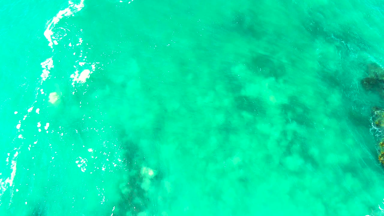 Overhead view of bright turquoise water at Buzzards Beach, Baja California Sur, with darker submerged rocks visible under the surface and no visible people.