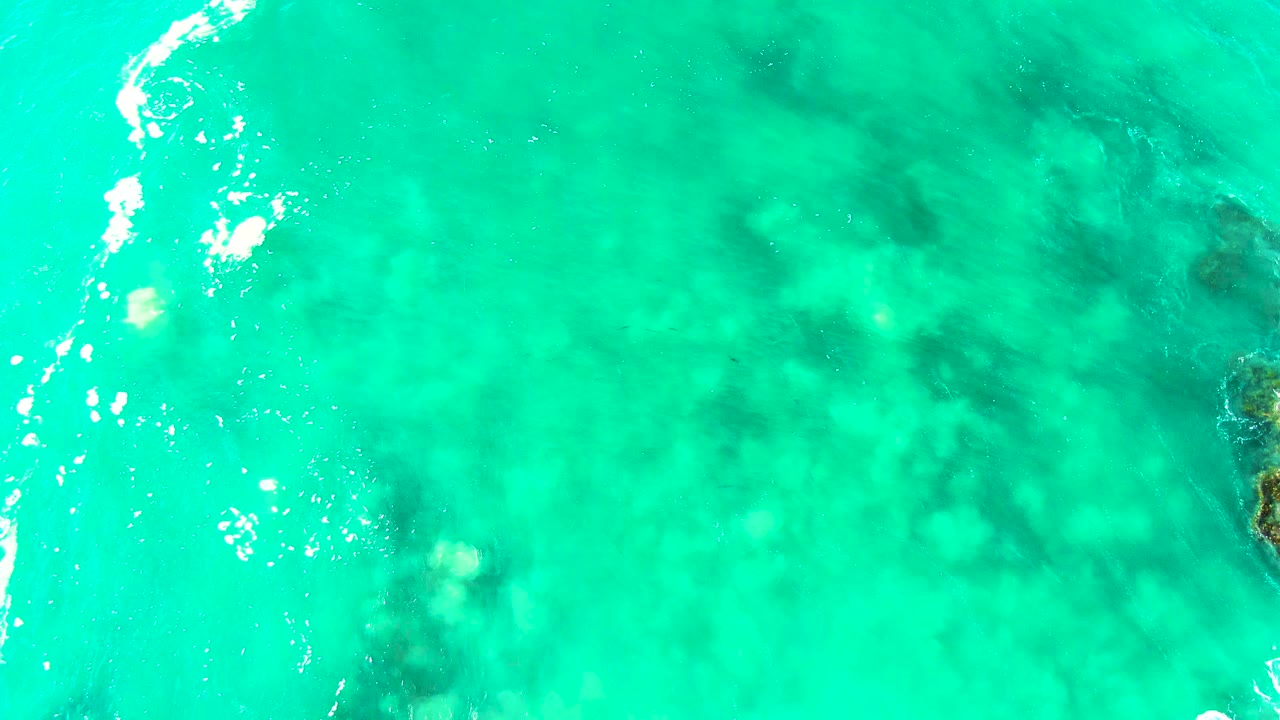 Clear turquoise ocean at Buzzards Beach, Baja California Sur, with a white foam line near the left edge and darker rock shapes beneath the water, no visible people.