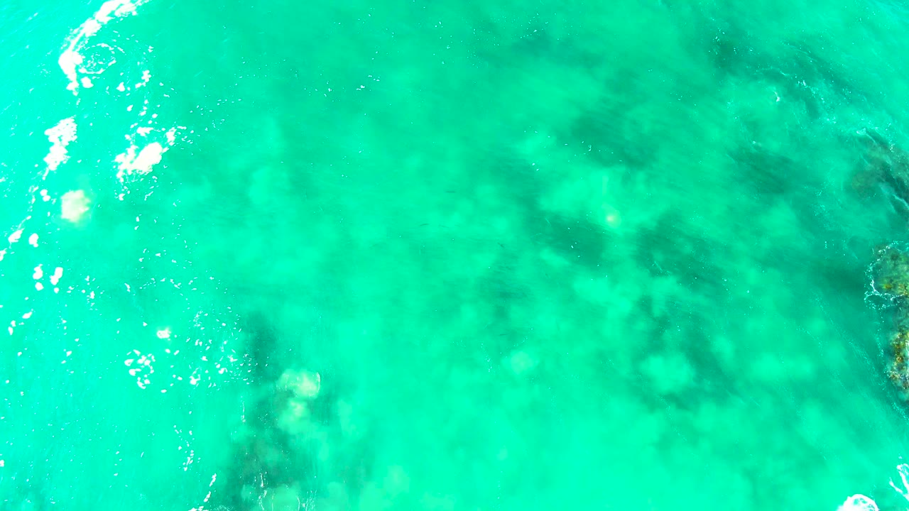 Transparent turquoise sea at Buzzards Beach, Baja California Sur, with mottled darker underwater formations and small bright foam highlights, no visible people.