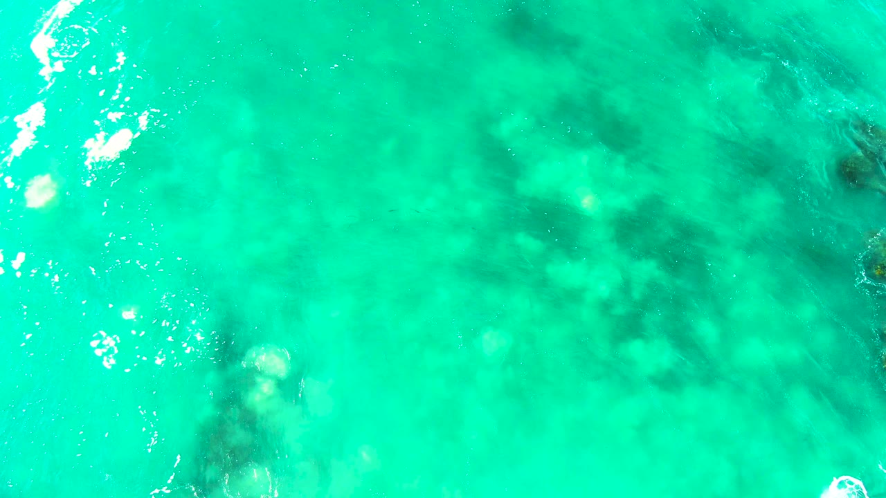 Overhead frame of shallow turquoise ocean at Buzzards Beach, Baja California Sur, with darker underwater patches shifted across the center and no visible people.