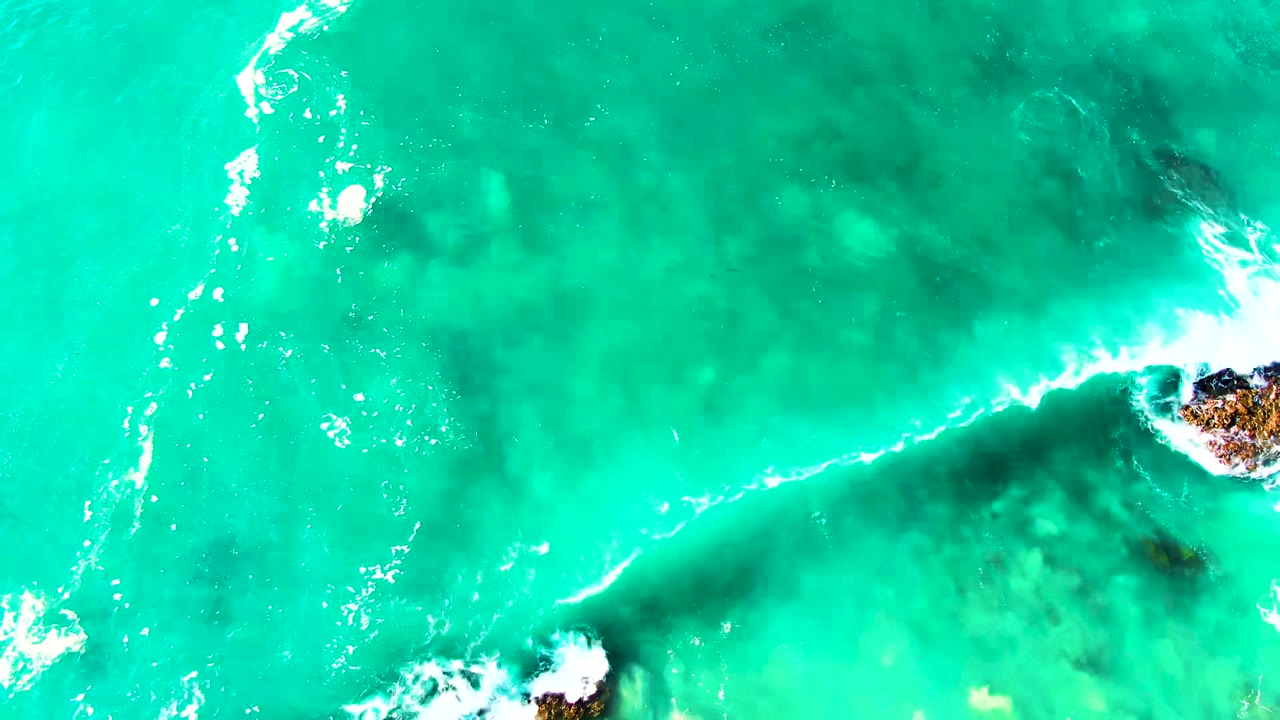 Top-down aerial frame at Punta Buzzards, Baja California Sur, showing turquoise water, white wave foam, dark submerged rock, small rocky outcrops at the edge, and no visible people.