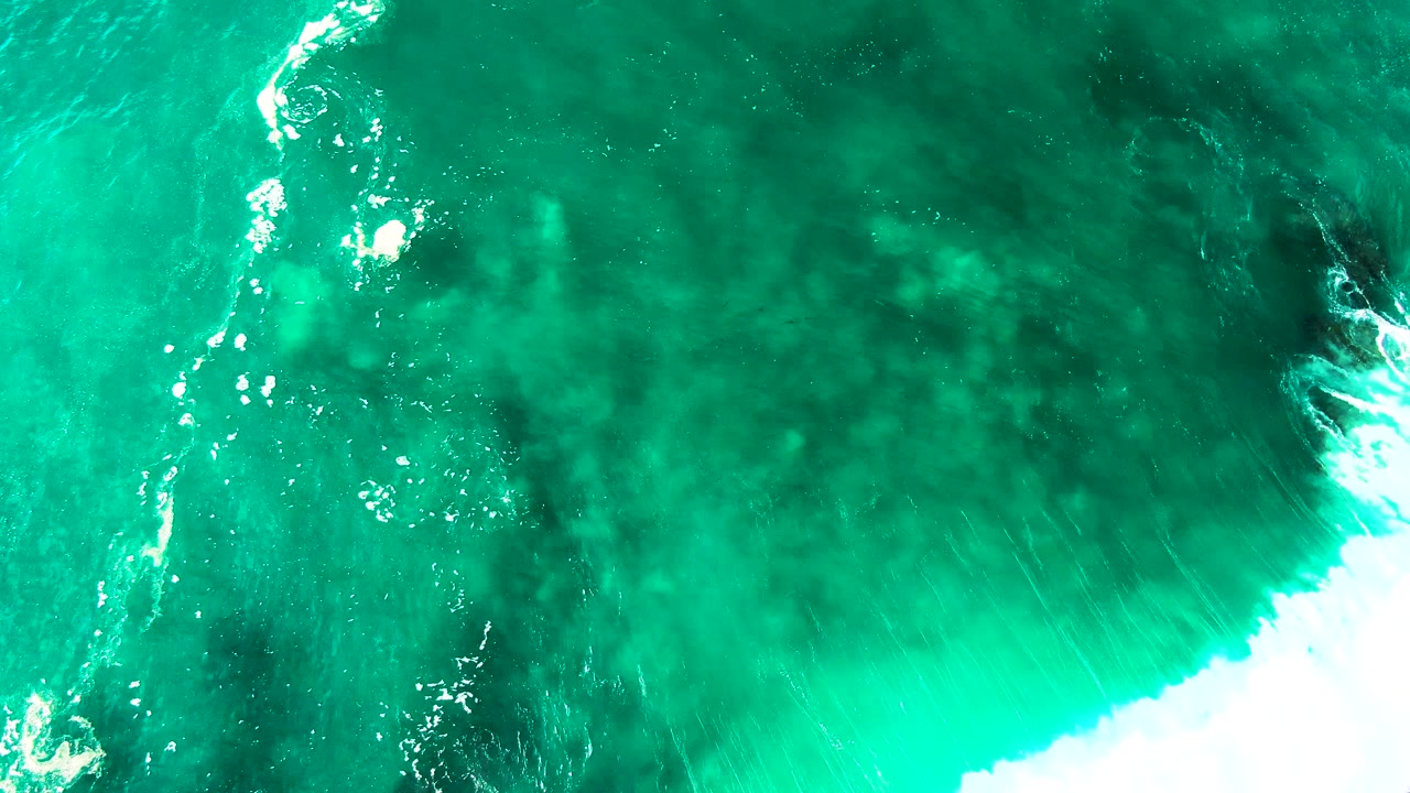 At 0:02, the aerial view over Punta Buzzards shows white surf sliding along the right side of turquoise water over darker seabed patches, with no visible people.