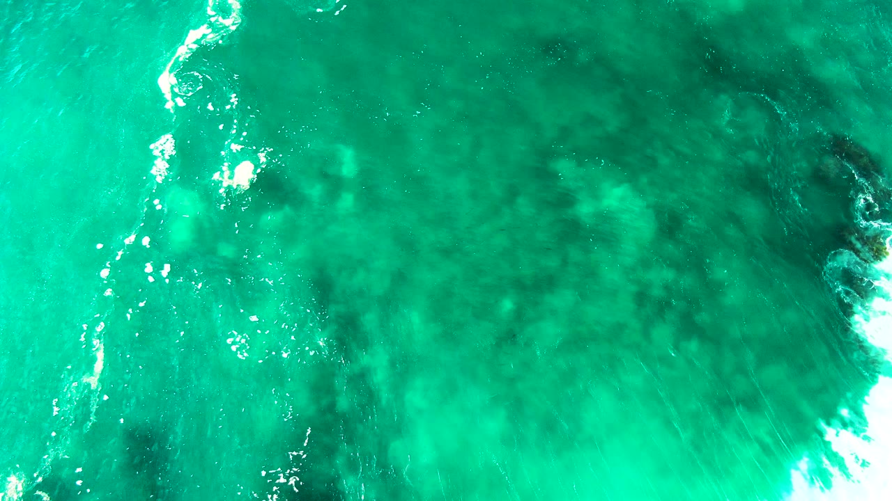 Aerial turquoise water with dark reef shapes and white foam at the frame edge.