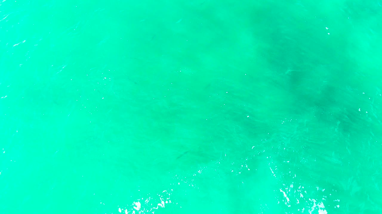 Bright turquoise water at Punta Buzzards shows scattered white foam highlights near the lower edge, darker shallow features, and no visible people.