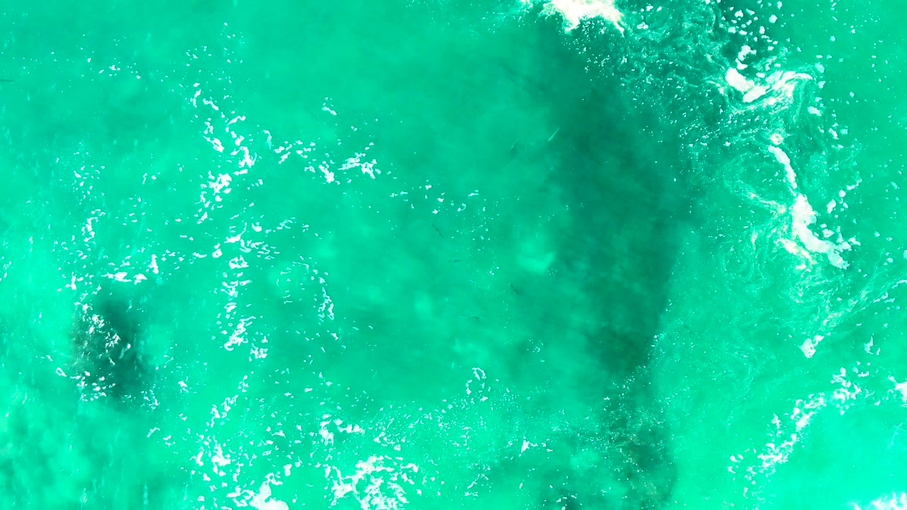 Bright turquoise ocean at Punta Buzzards shows a longer dark submerged formation through the water, small foam streaks, and no visible people.