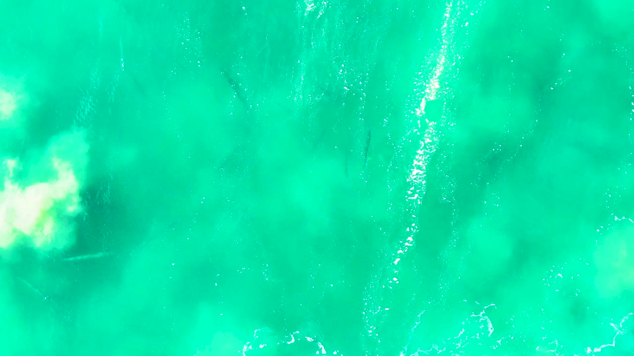 Aerial turquoise ocean surface at Punta Buzzards with a bright white foam patch near center left, faint fish shapes under the water, and no visible people.