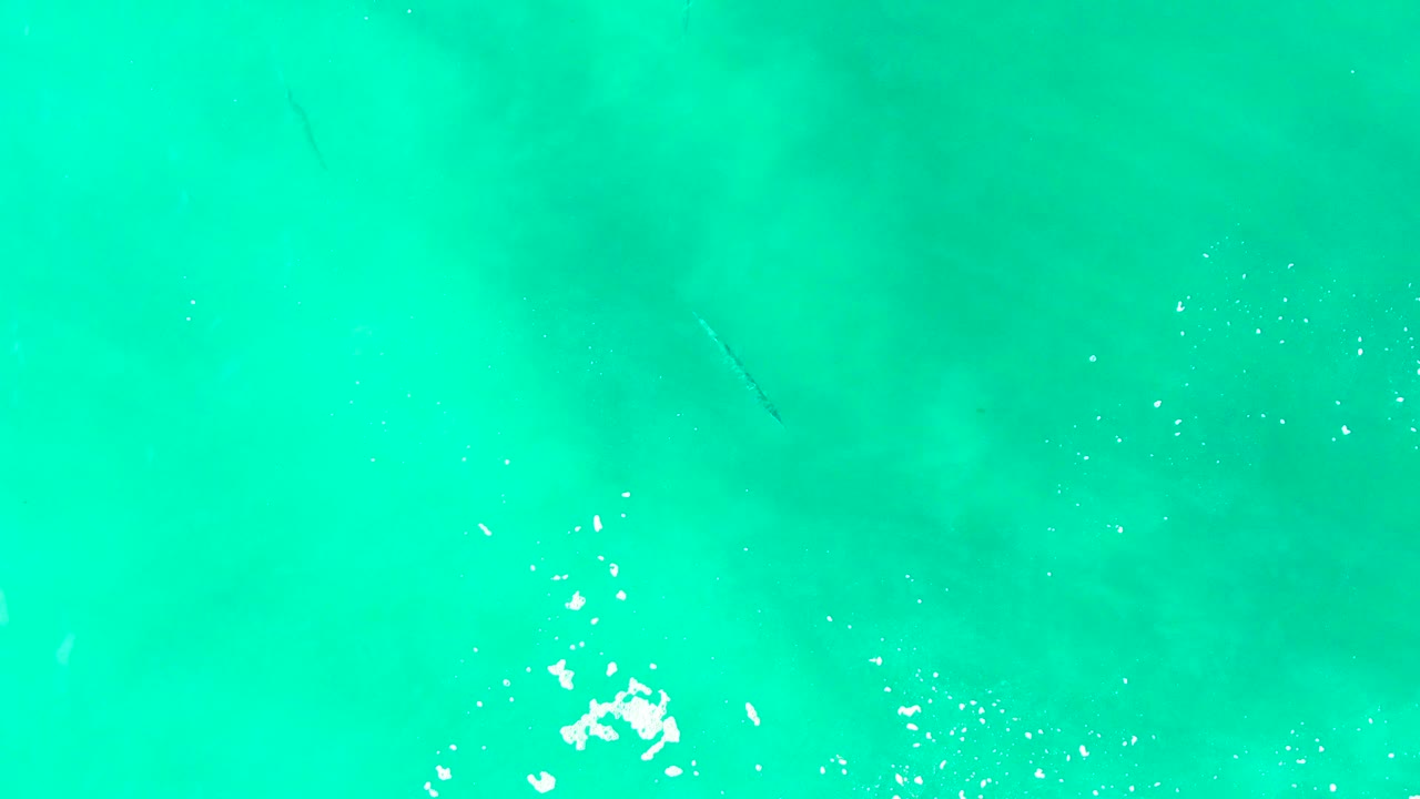 Turquoise water at Punta Buzzards with a faint fish shape near the center area and bright white splash marks along the lower edge, no visible people.