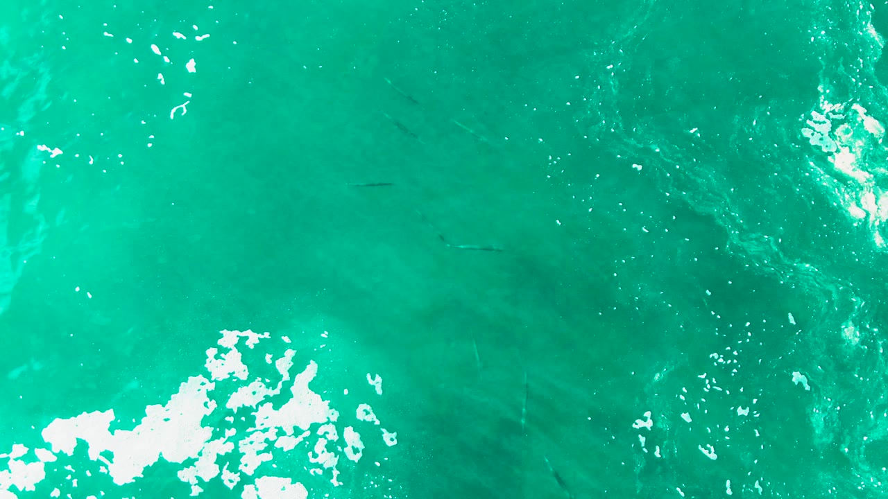 Overhead turquoise ocean at Punta Buzzards with bright white foam along the lower edge, sun glints on the water, and no visible people.