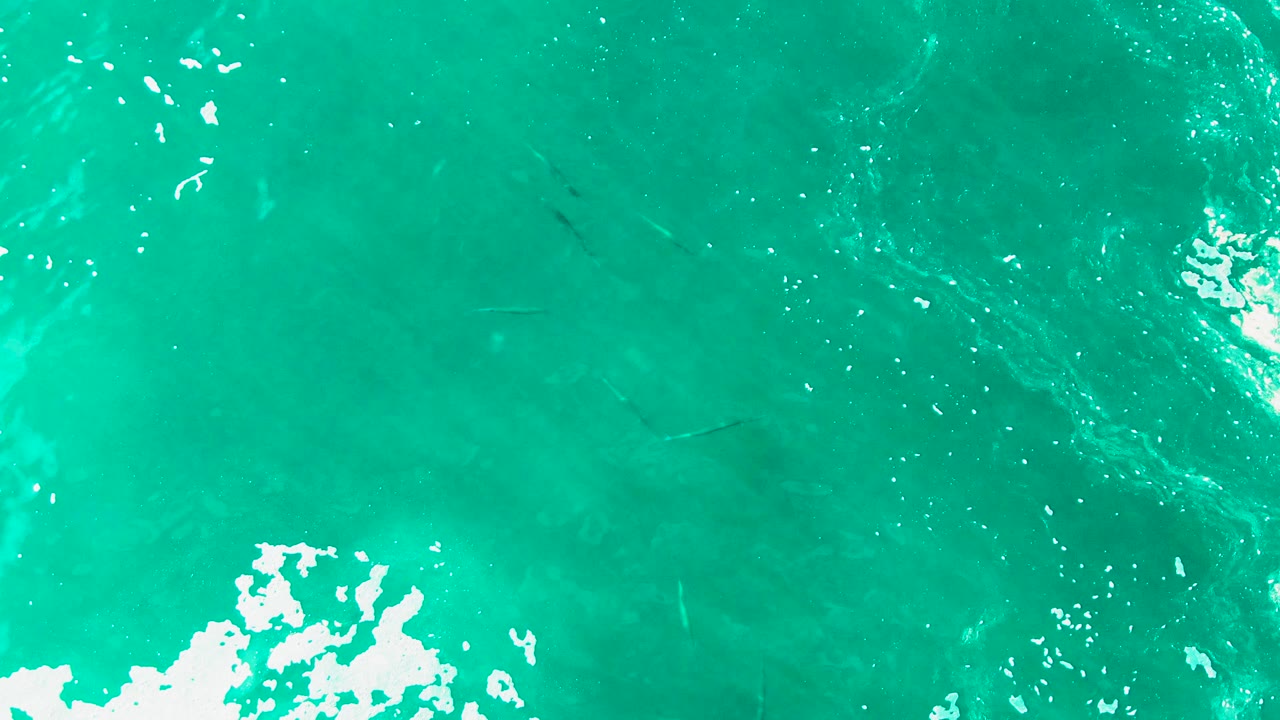Clear turquoise ocean at Punta Buzzards seen from above, with a larger white breaking foam patch at the bottom and no visible people.