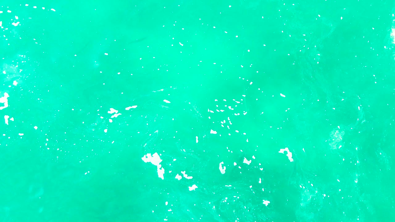 Bright turquoise ocean at Punta Buzzards with a small white splash cluster left of center and faint darker water texture below, with no visible people.