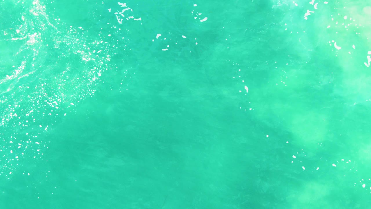 Overhead frame of clear blue-green ocean at Punta Buzzards with bright sparkle across the upper center, pale underwater sand tones, and no visible people.
