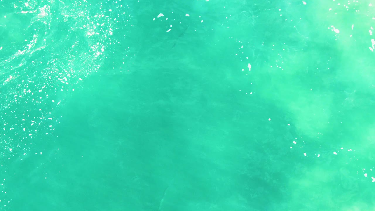 Straight-down view of turquoise ocean water in Baja California Sur, with soft pale shapes below the surface and small white highlights near the top edge, no visible people.