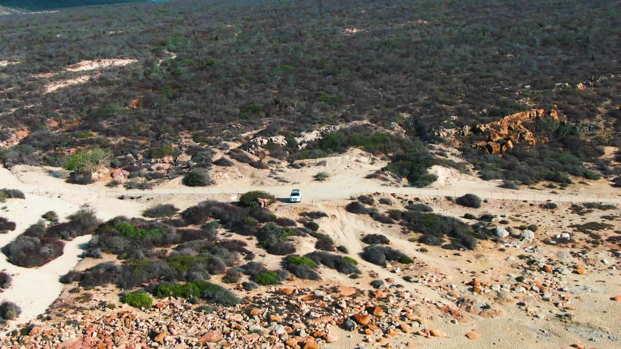 Aerial view of a white car on a pale dirt road through rocky scrub near Buzzards Beach East Cape, Baja California Sur, with no visible people.