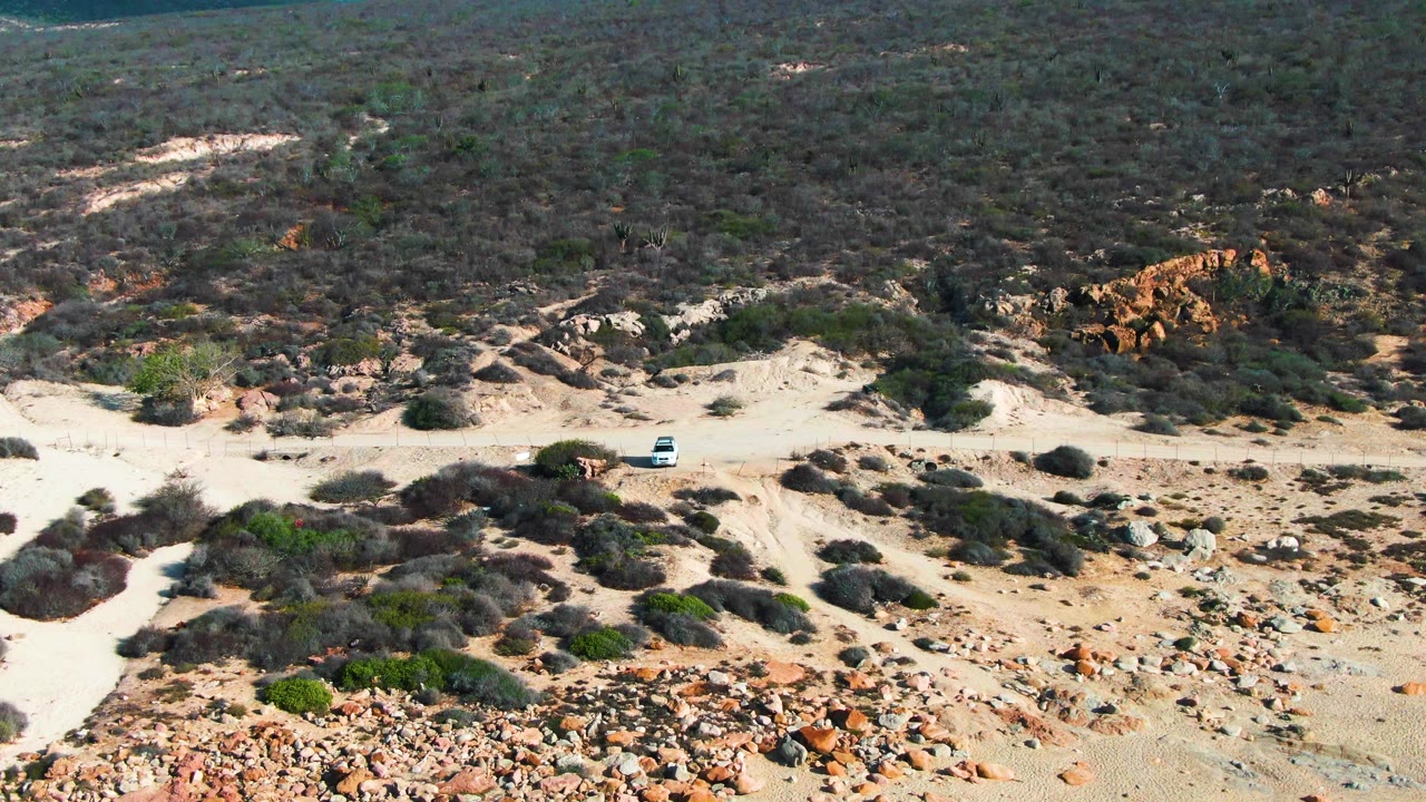 Aerial frame of the white car slightly farther along the dirt track in the arid Buzzards Beach East Cape landscape, with no visible people.