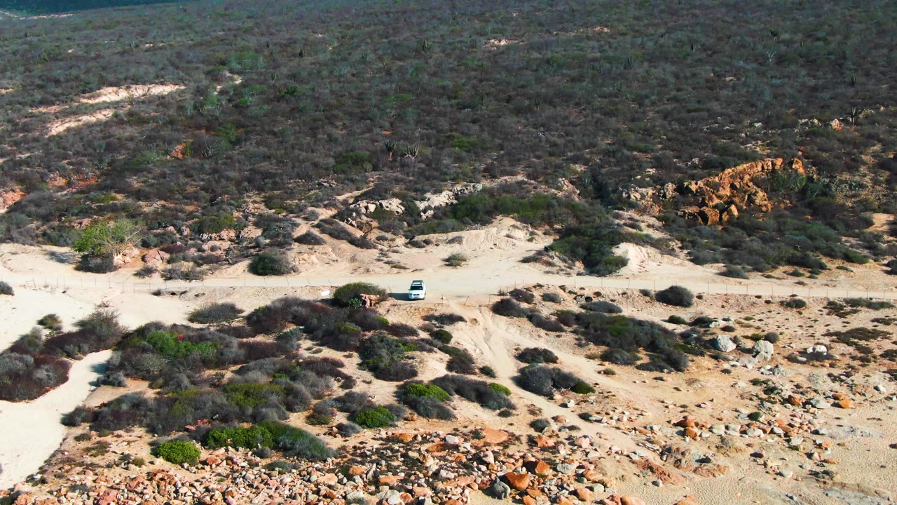 The white vehicle remains on the sandy road beneath darker scrubland and rust-colored rocks in Punta Buzzards, Baja California Sur, with no visible people.