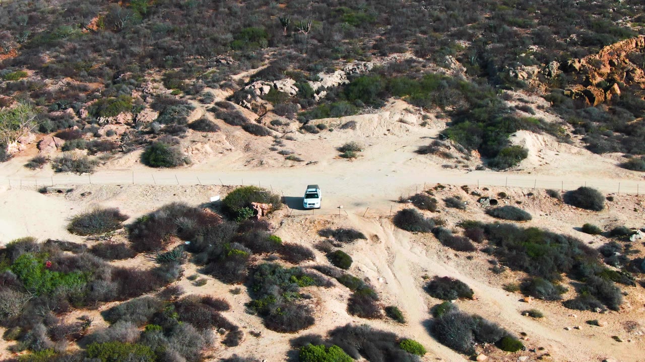 At Buzzards Beach East Cape in Baja California Sur, a white car appears on a pale dirt road below dry shrubs and eroded sandy slopes, with no visible people.