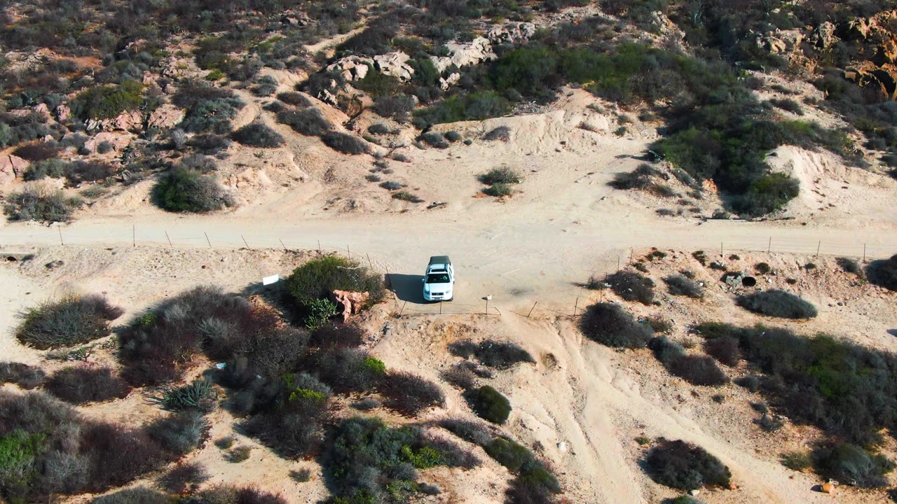 A high aerial view shows the white car on the dirt road near small eroded gullies and scattered shrubs in Baja California Sur, with no visible people.
