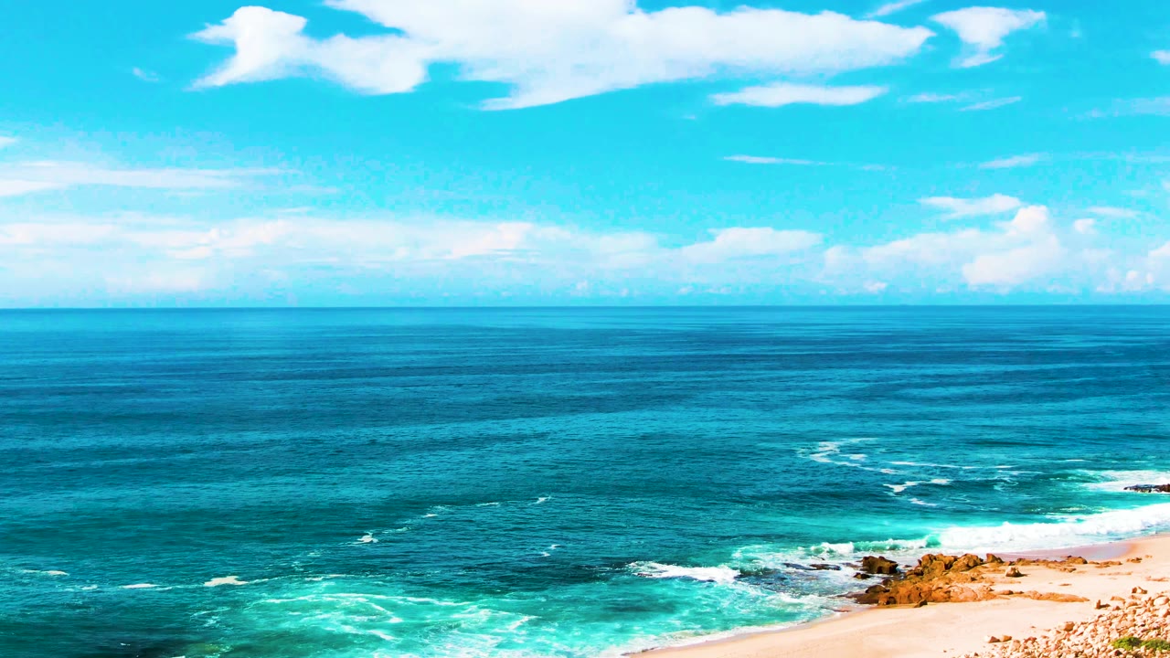 Punta Buzzards shore appears at the lower right as white waves break around rocks below a wide blue ocean and scattered daylight clouds, with no visible people.
