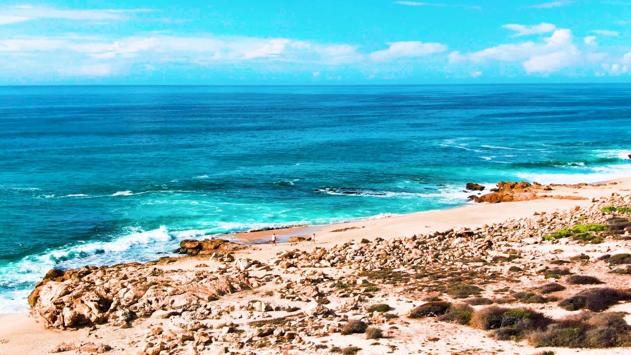 Elevated view of Punta Buzzards beach in Baja California Sur with turquoise ocean, white surf, tan rocks, low scrub, pale sand, and a small person walking near the shoreline.