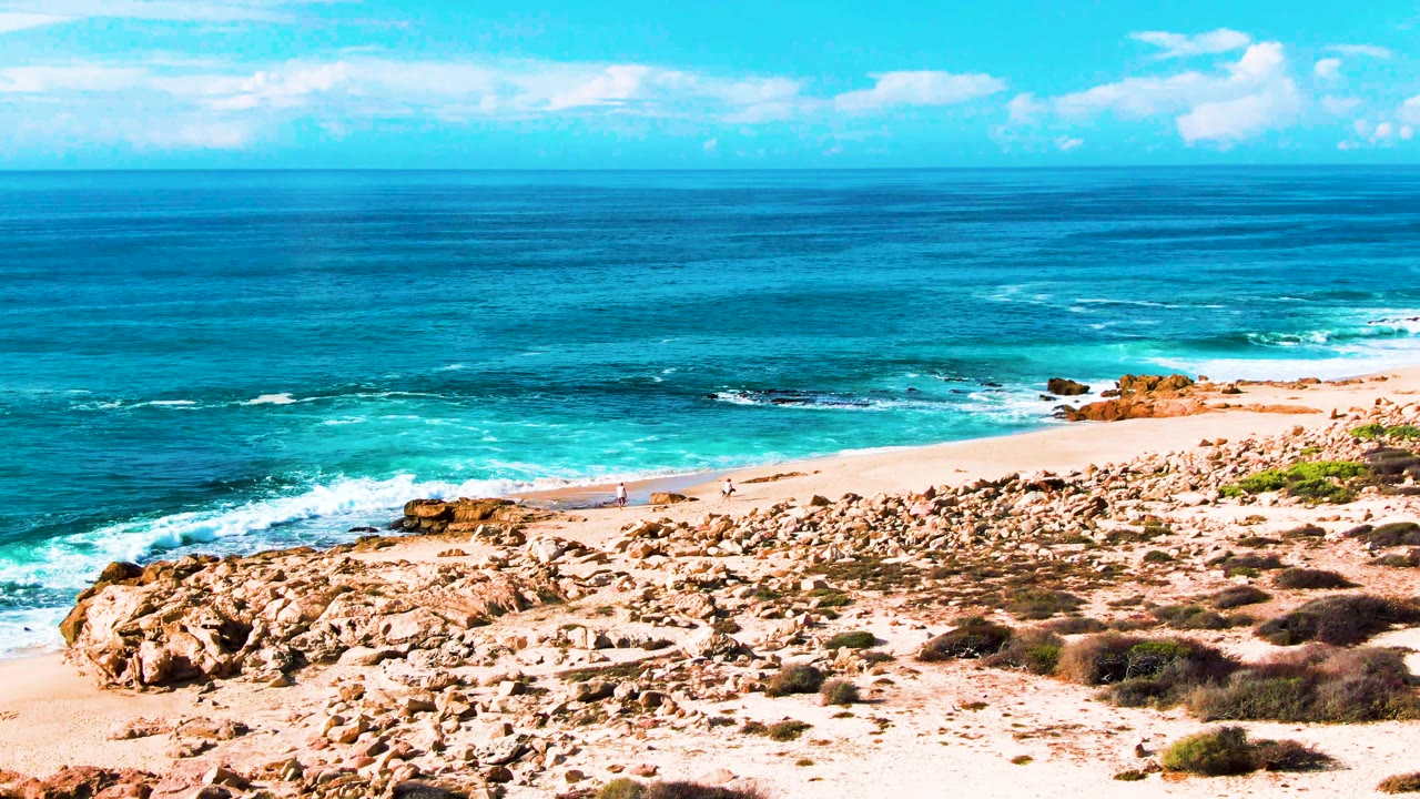 Rocky beach foreground with blue ocean, breaking waves, and a tiny walker on the sand.