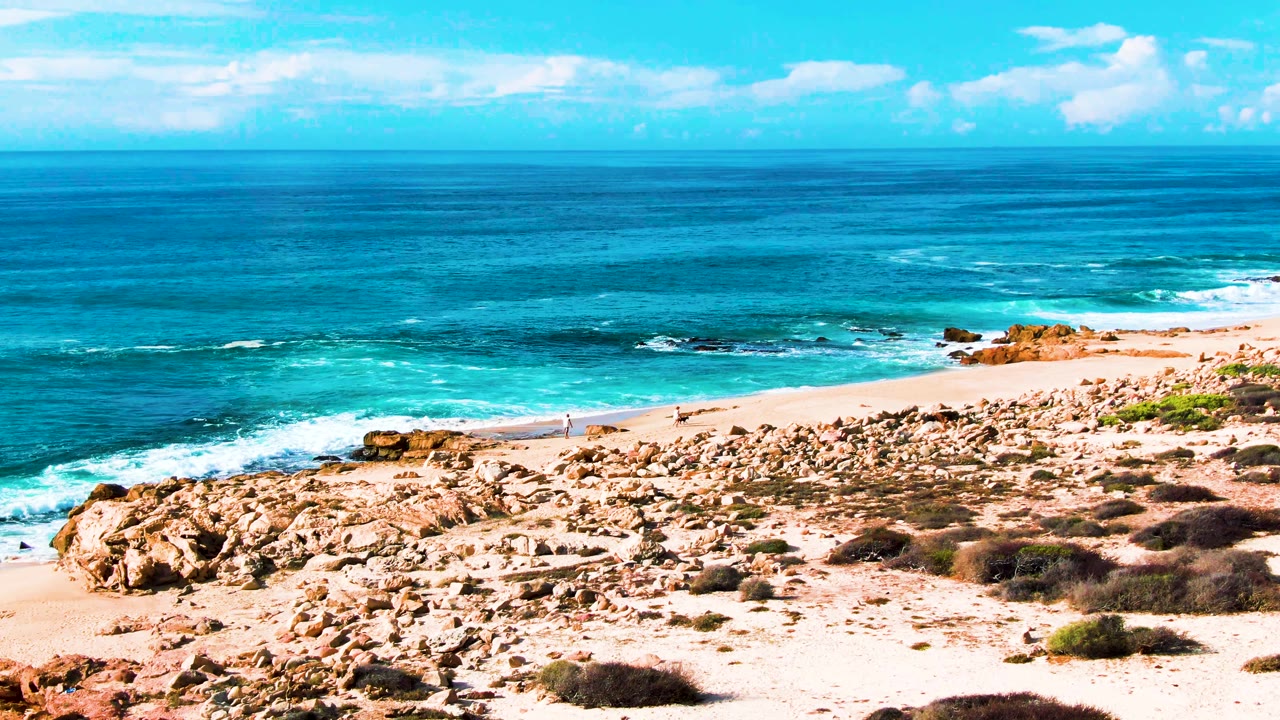 Wide elevated view at Punta Buzzards showing layered blue ocean, white wave bands, a sandy beach bordered by rocks and scrub, and a distant person walking near the waterline.