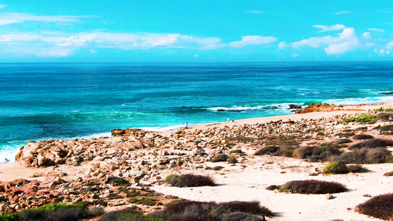Wide view of Buzzards Beach in Baja California Sur with turquoise ocean, rocky sandy shore, foreground scrub, scattered clouds, and no visible people.