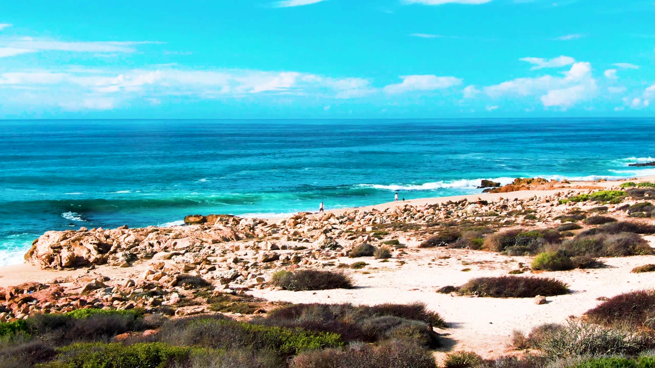 Wide East Cape coastal view at Buzzards Beach with rocks across the midground, sandy patches, scrub vegetation, blue ocean, and no visible people.