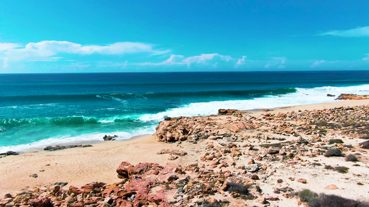 A line of white foam breaks near the Punta Buzzards sandy shore while reddish rocks fill the lower right foreground and no visible people are present.