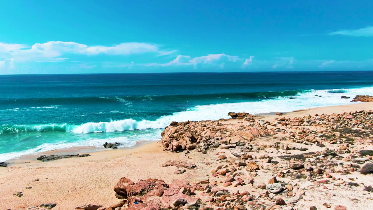 The surf line shifts closer to the sandy beach at Punta Buzzards, with dark rocks near the waterline, tan rock ledges in the foreground, and no visible people.
