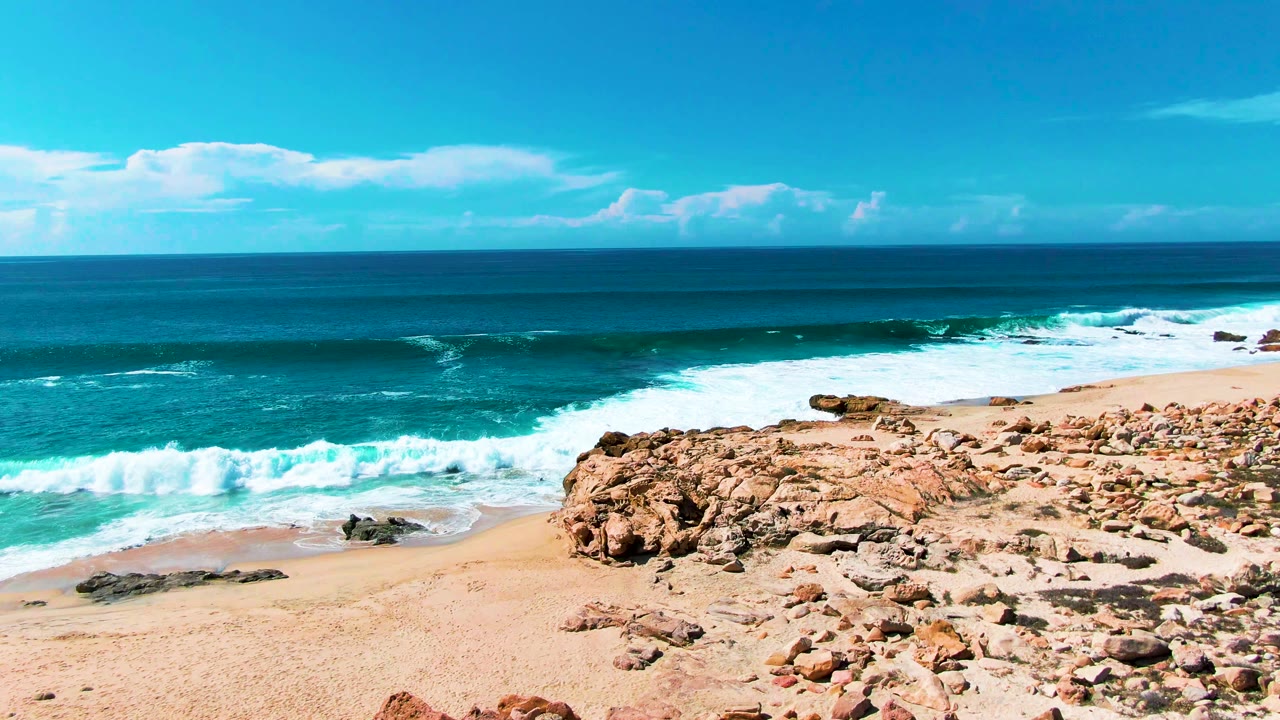 White surf spreads across the sand below the rocky Punta Buzzards coastline, with layered blue ocean beyond the break and no visible people in frame.