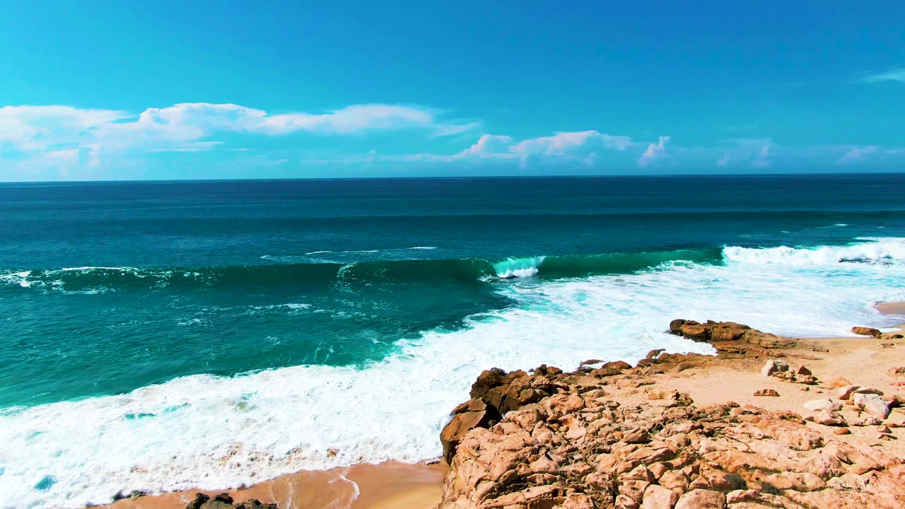 Wide ocean surf breaking beside tan rocks and sandy beach