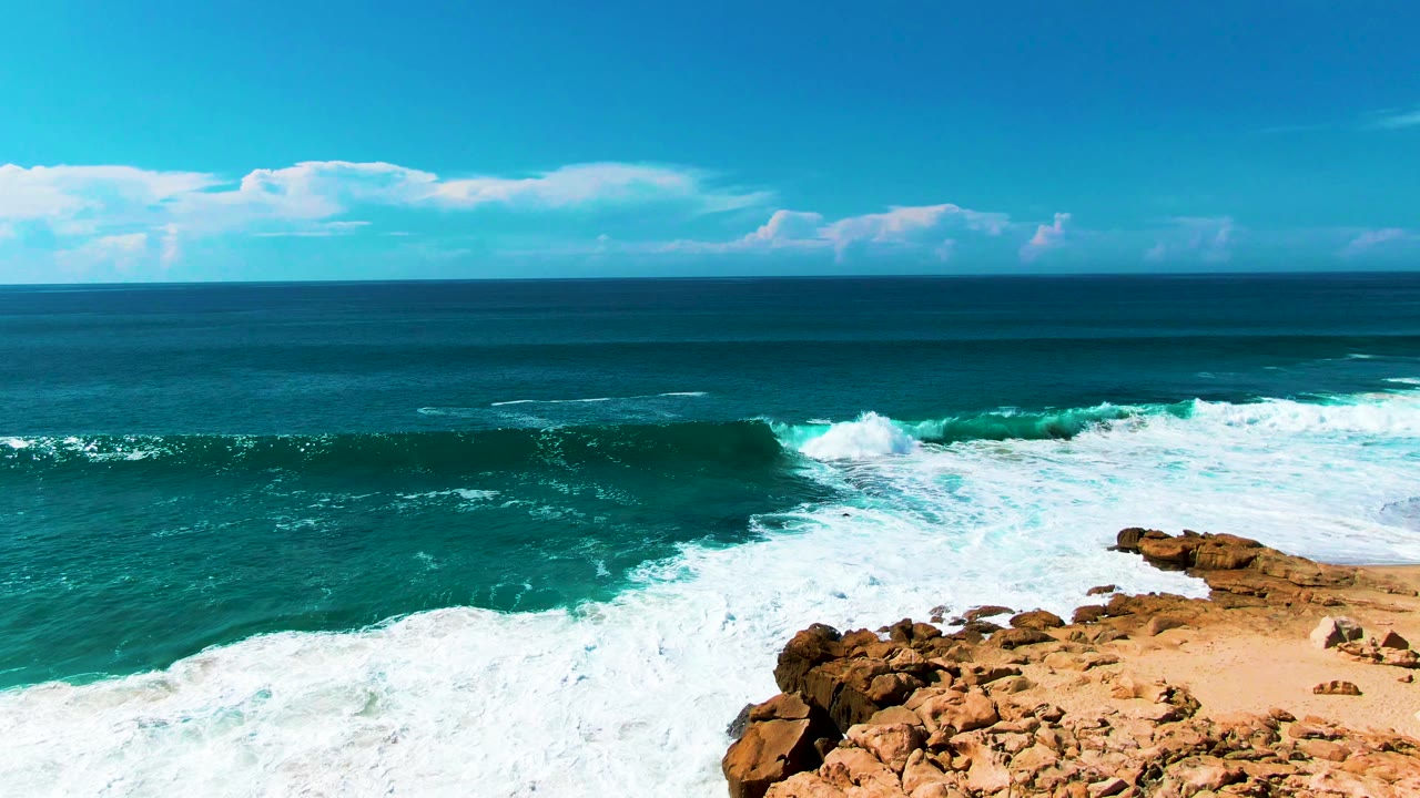 A larger band of whitewater breaks across the turquoise water near the Punta Buzzards rocks, with the horizon and scattered clouds steady in the background and no visible people.