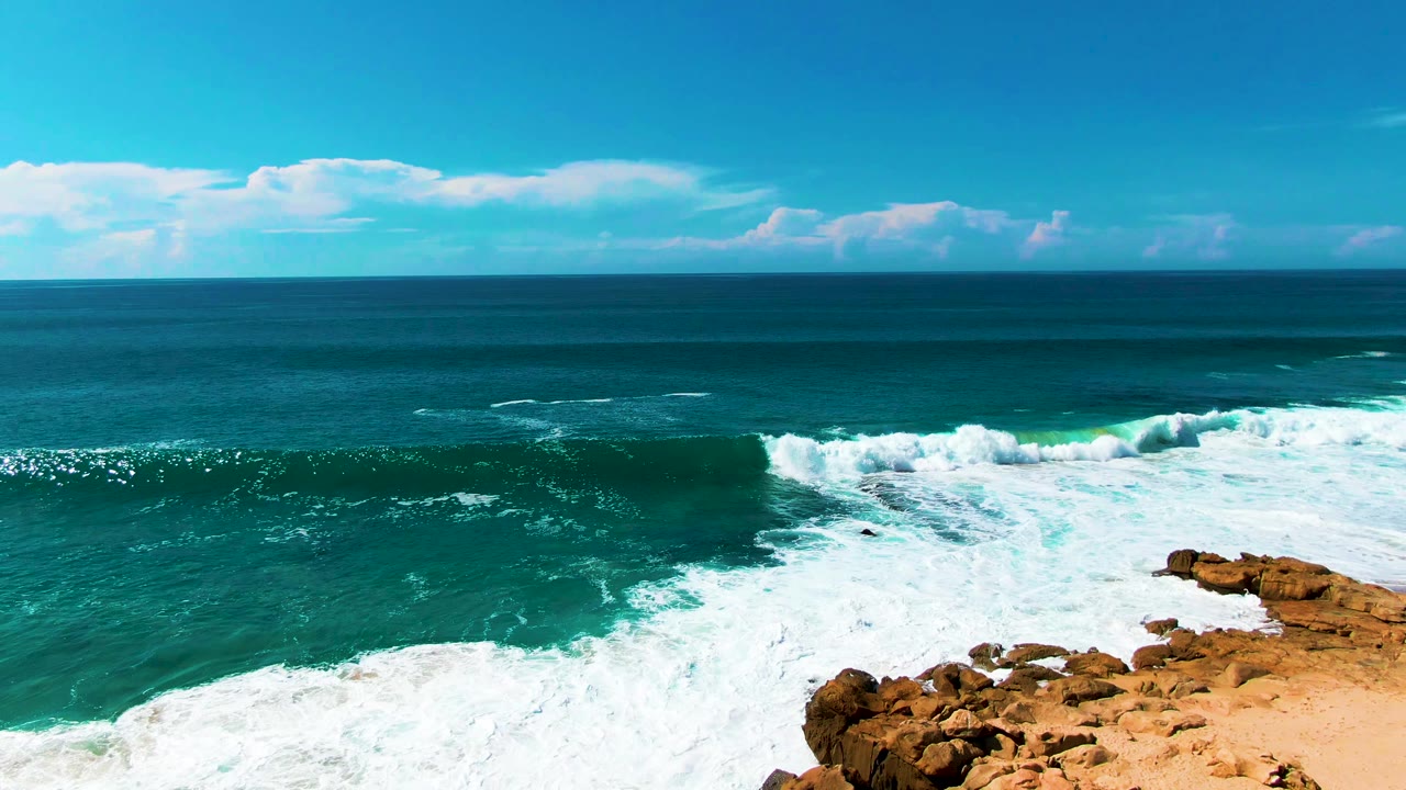 White surf curls around the rocky point at Buzzards Beach while deep blue water fills the horizon line and tan rocks remain on the lower right, with no visible people.