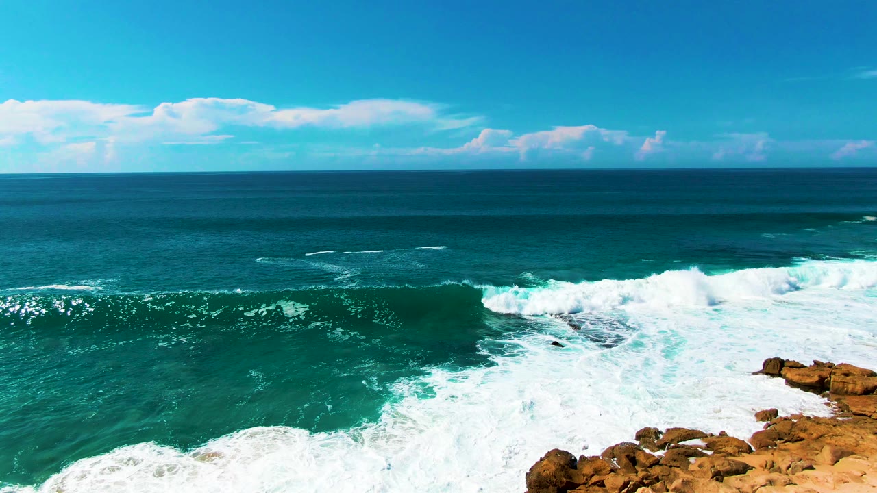 A broad sheet of white foam covers the nearshore water beside exposed tan rocks at Punta Buzzards, with open ocean and cloud bands beyond and no visible people.