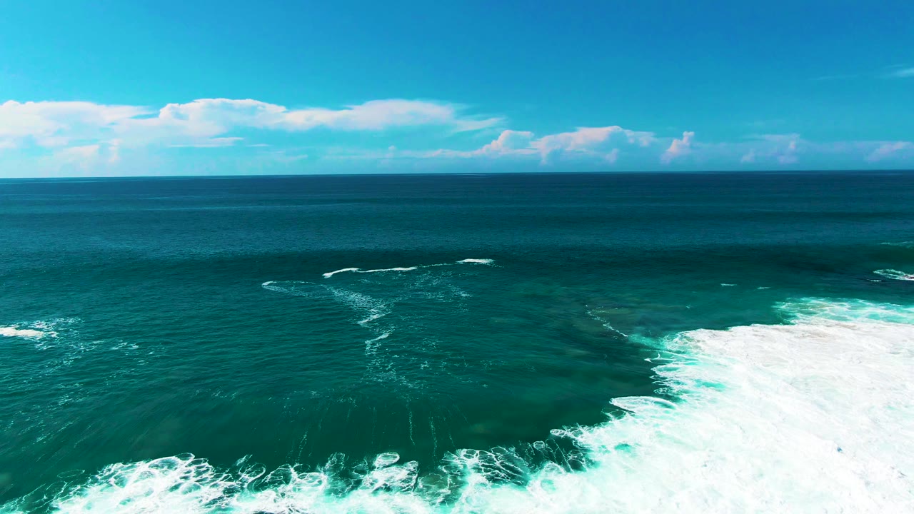 Aerial ocean view at Punta Buzzards, Baja California Sur, as white foam thins along the lower edge while teal water and a clouded horizon remain visible, with no visible people.