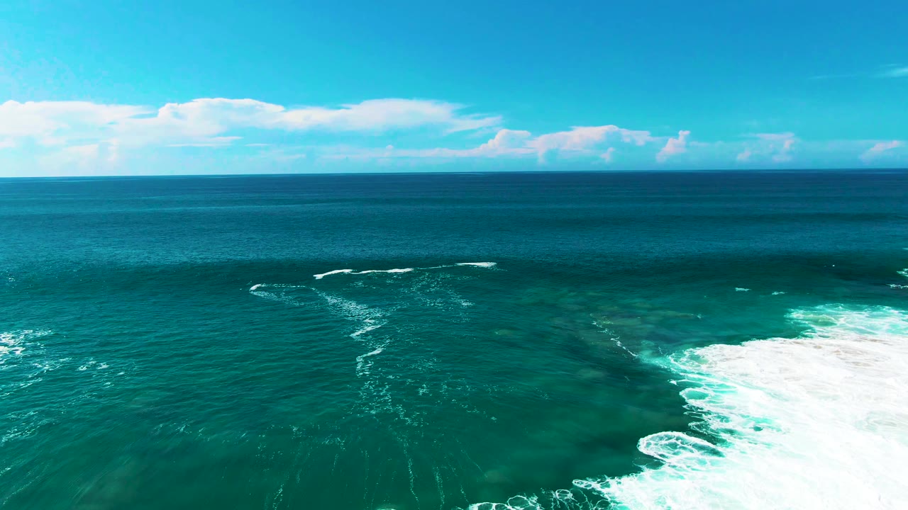 Punta Buzzards aerial frame with reef-toned shallows beneath turquoise water, a patch of white surf foam at the lower right, and no visible people.