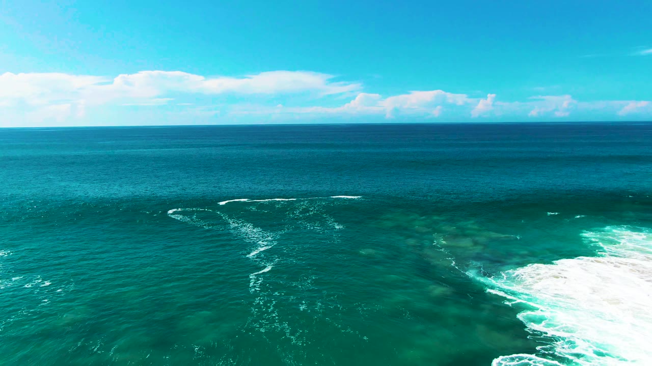 Wide turquoise sea under scattered clouds with surf foam mostly at the far right edge.