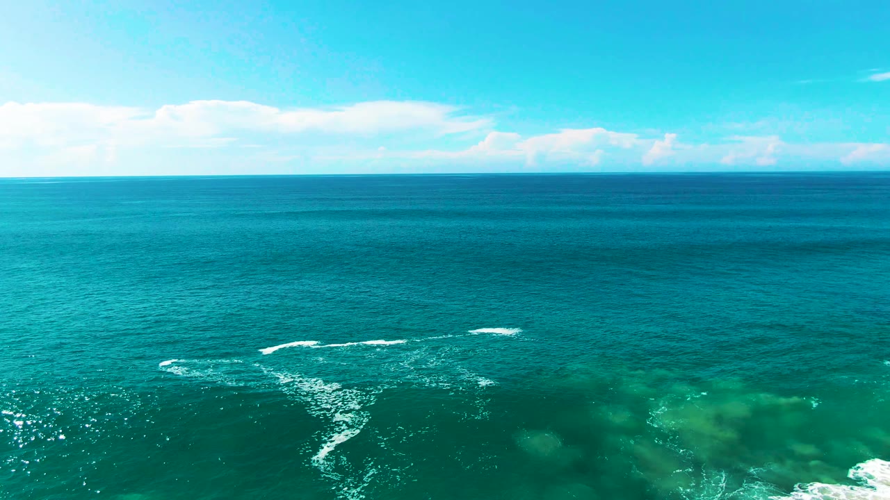 Aerial view over Punta Buzzards turquoise ocean with a distant horizon, pale clouds, small white wave breaks, and no visible people.