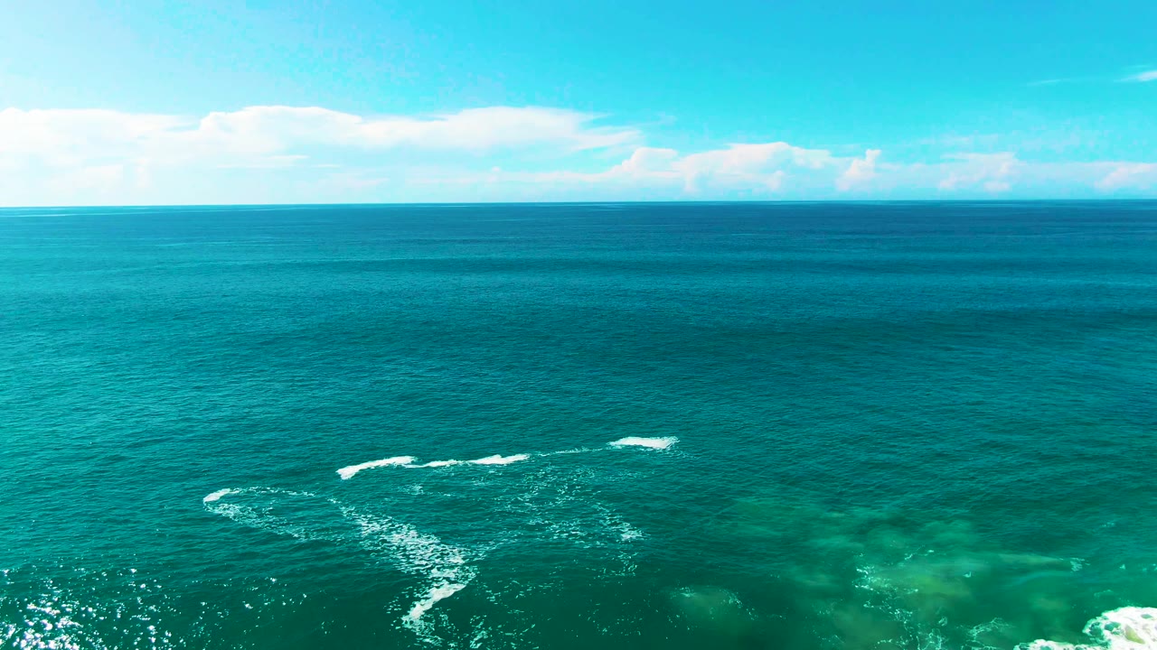 Wide aerial frame from Punta Buzzards showing turquoise and deeper blue water, faint seabed patches, low clouds near the horizon, and no visible people.