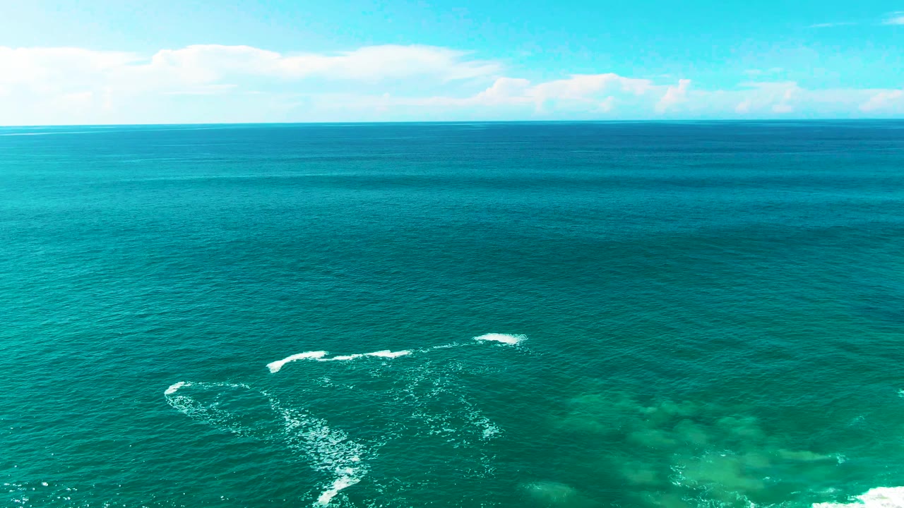 Punta Buzzards aerial ocean frame with the horizon high in view, scattered cloud shapes, whitecaps across the center, green-blue shallows, and no visible people.