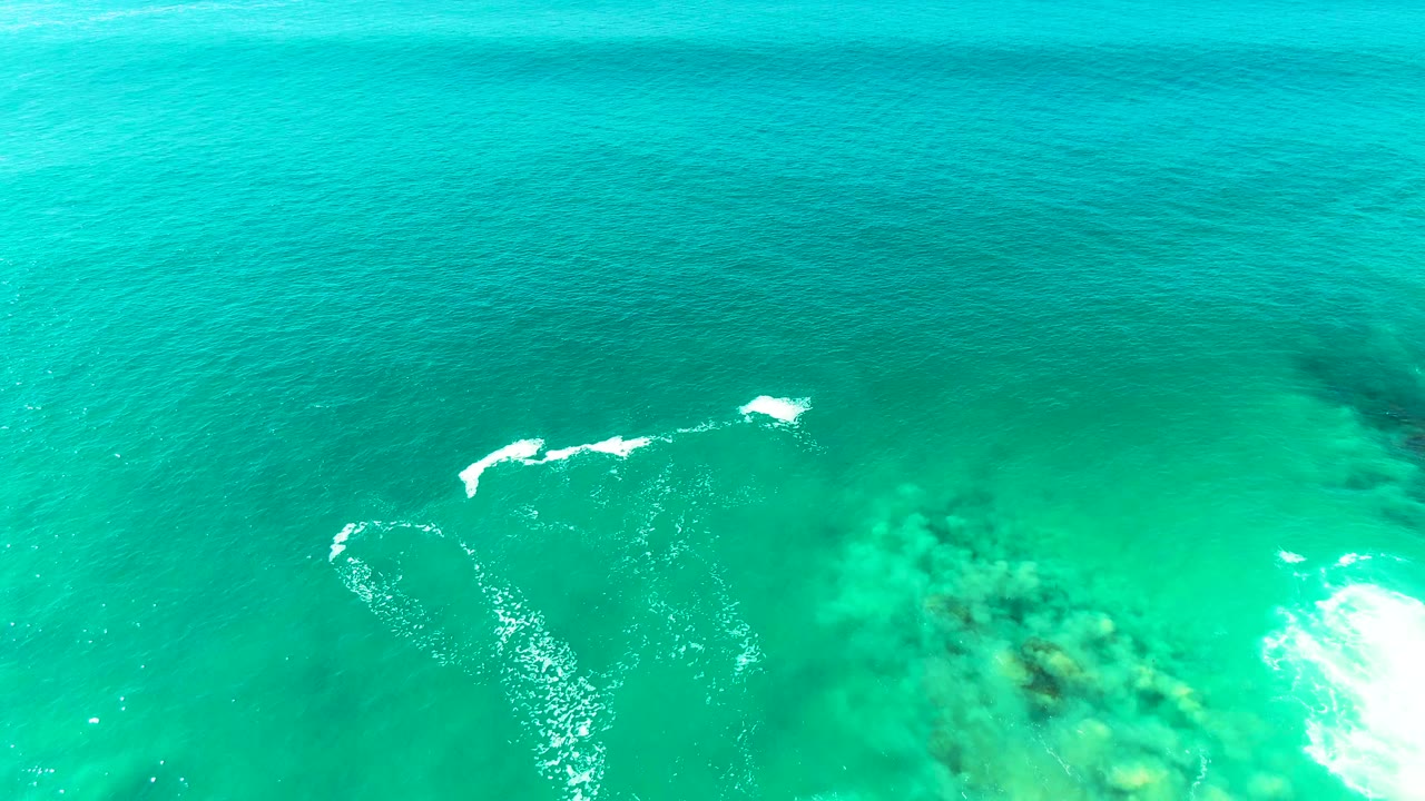 Aerial view over turquoise water at Punta Buzzards, Baja California Sur, with white wave foam near submerged rock or coral and no visible people.