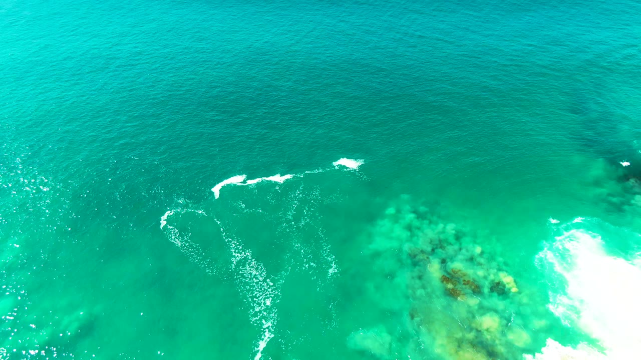 Top-down view at Punta Buzzards with bright white breaking foam on the right and reef-like submerged forms visible through turquoise water, with no visible people.