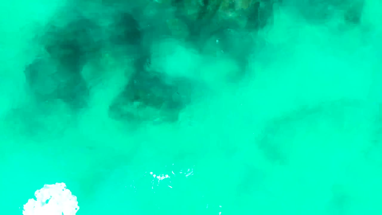 Top-down view of turquoise water at Punta Buzzards, Baja California Sur, with dark underwater rock or coral shapes near the upper frame, a white foam patch at the bottom edge, and no visible people.