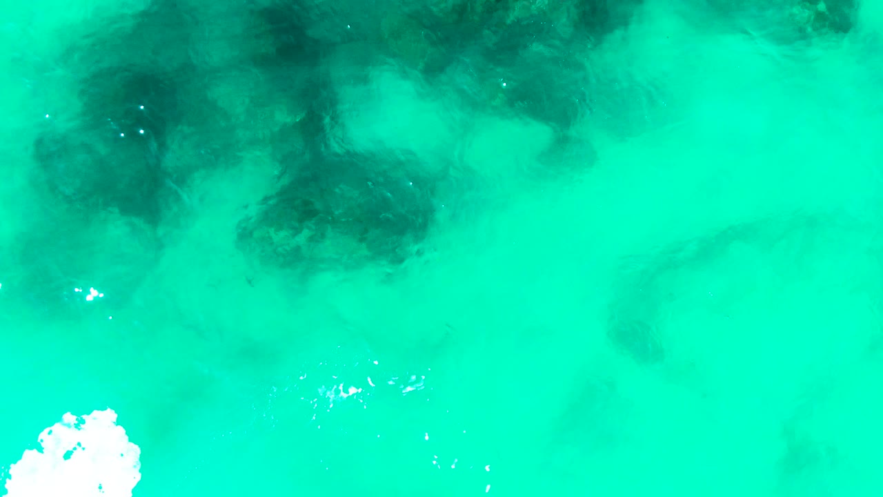 Aerial frame over Punta Buzzards turquoise water with darker submerged formations across the upper half, bright white foam near the lower center, small surface glints, and no visible people.