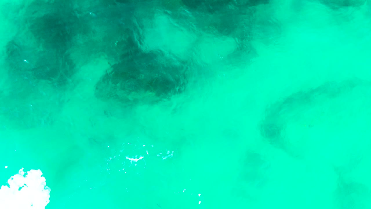 Top-down Punta Buzzards water view showing dark rock or coral patches beneath green-blue water, with a bright foam shape pushing up from the lower edge and no visible people.