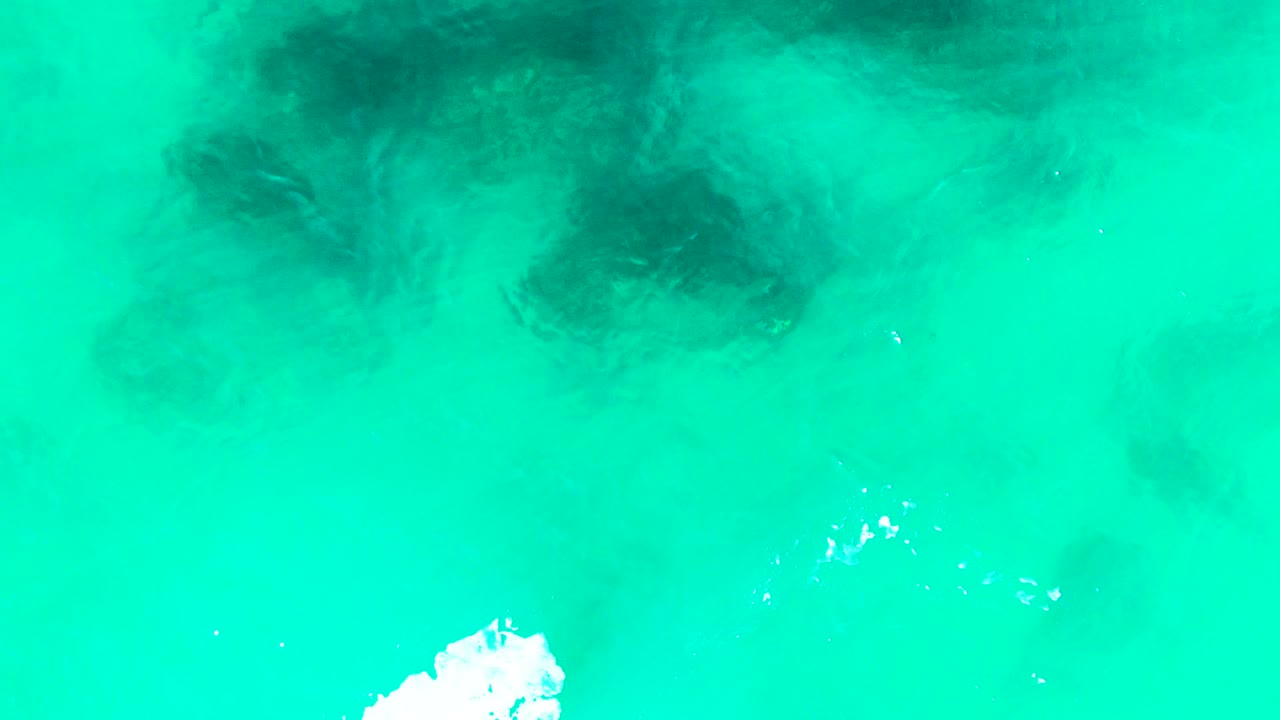 Aerial view at Punta Buzzards showing bright turquoise water, a dark underwater patch near the upper center, faint sandbar gradients, a small foam mark below, and no visible people.